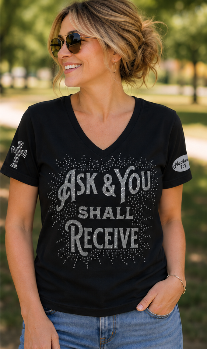 A woman wearing a black V-neck t-shirt with silver rhinestone text that reads 'ASK & YOU SHALL RECEIVE' on the front, along with a cross graphic on the right sleeve and a small logo patch on the left sleeve, photographed outdoors in a park setting.